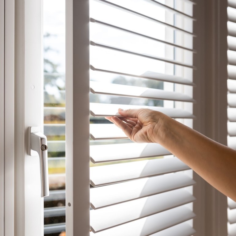 Close-up of a hand adjusting white plantation-style window shutters to control light and privacy in a modern home.
