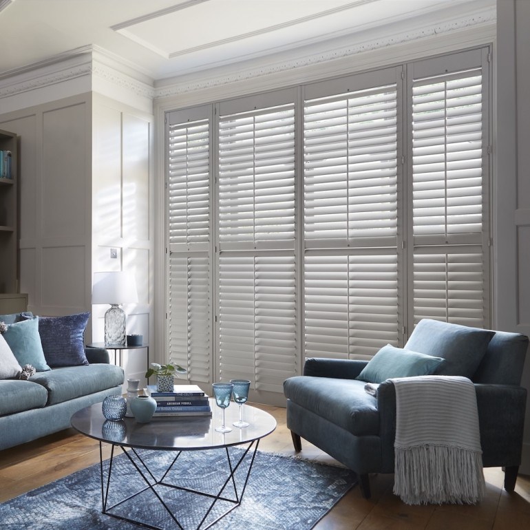 Stylish living room with white plantation shutters on tall windows, filtering natural light and complementing a coastal-inspired interior.