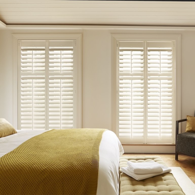 Bright bedroom with white full-height shutters on tall windows, offering light control and privacy while complementing a warm, modern interior.
