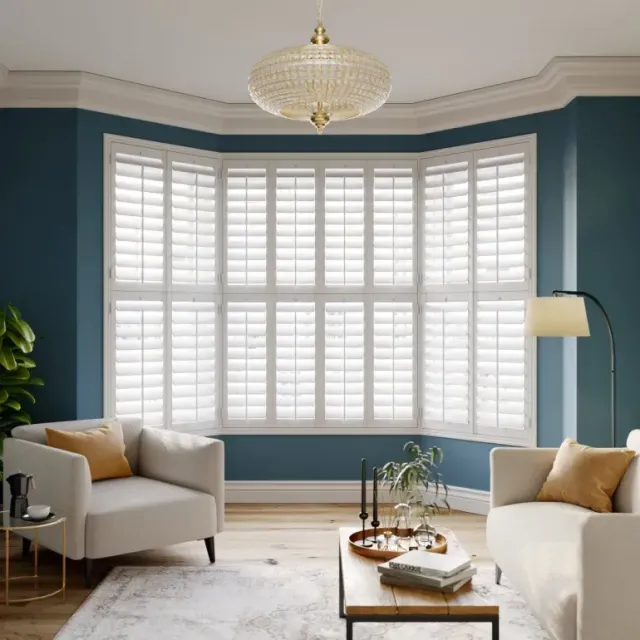 Elegant tier-on-tier white window shutters fitted in a bay window of a modern living room with blue walls and neutral furniture.