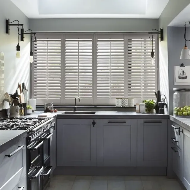 Modern kitchen with grey cabinetry and full-height white shutters on large windows, offering light control and timeless style.